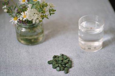 Parlak yeşil spirulina hapları, bir bardak su ve çiçekli vazo. Vejetaryenler için sağlıklı gıda desteği. Günlük rutinde demirin doğal kaynağı. Bitkisel anemi tedavisi. Ferritin eksikliği olan insanlar için ilaçlar.