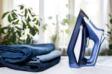 Mavi elbise ütüsü ve arka plandaki ütü masasında bir yığın kot pantolon. Ev İşleri Ekipmanı