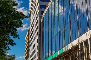 Ticari binanın dış cephesi ve mavi gökyüzüne karşı yeşil ağaç. İnşaat sırasında camları camlı olan konut, otel, hastane veya diğer ticari tesisler..
