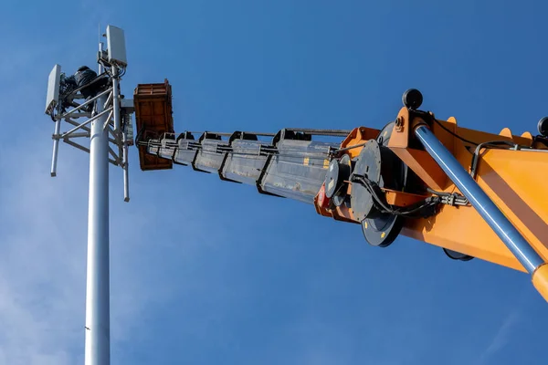 Crane with telescopic boom lift used as an aerial working platform. Worker install cellular base station with transmitters 3G, 4G and antennas on cell tower on background of blue sky. Focus on arrow.