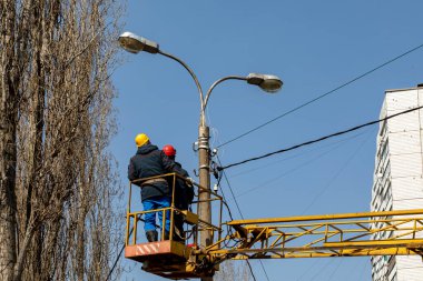 Kasklı ve korumalı ekipmanlı belediye çalışanları sokak lambasına kablo döşüyorlar.