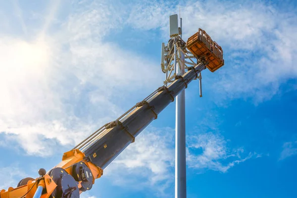 Crane with telescopic boom lift used as an aerial working platform. Worker install cellular base station with transmitters 3G, 4G, and antennas on cell tower on blue sky background.