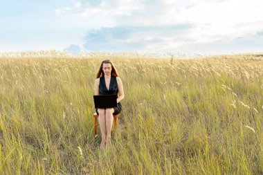 Ofis elbiseli bir kadın, sahada tahta bir sandalyeye oturur ve dizüstü bilgisayarla çalışır. Serbest konsept, iş dengesi ve dinlenme.