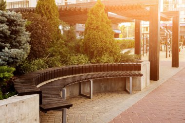 Gün batımında küçük bir şehir bahçesinde yarı dairesel ahşap bir bank. Pergola, çalılar ve selvi ağaçları arka planda