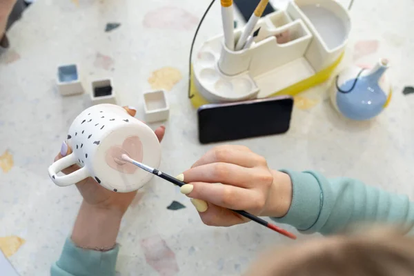 Womens hands paint pink heart on back of ceramic cup with brush. Training in profession of ceramicist, improvement of skills. Ceramic art concept.