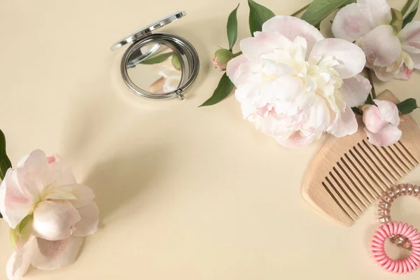 Hairbrush or comb, small mirror, hair spirals and large peony buds on a beige background. The concept of female beauty. Hair care at home. Copy space