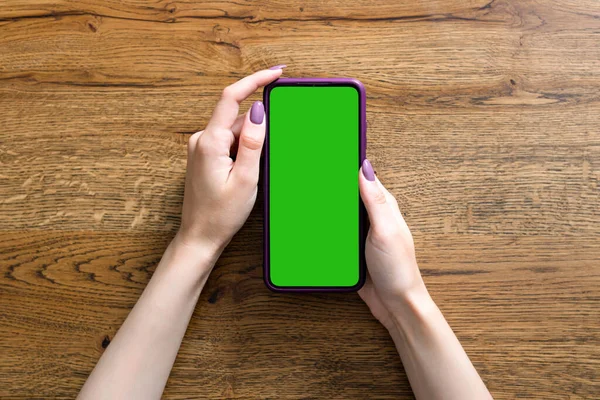 Beautiful female hands hold a smartphone on wooden background. Empty green screen for inserting your own picture or text. — Stock Photo, Image