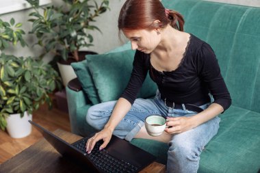 Bilgisayarda çalışan ve kahve içen çekici genç bir kadın. Dizüstü bilgisayar kullanmak ya da evde çalışmak.