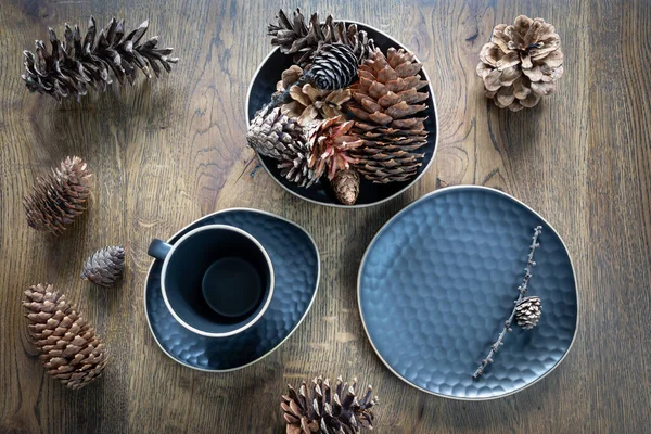 Conjunto de platos azul oscuro: taza de té, platillo, plato, un tazón con conos de abeto, variedad de conos sobre fondo de madera. Decoración de mesa de Navidad, vacaciones mágicas — Foto de Stock