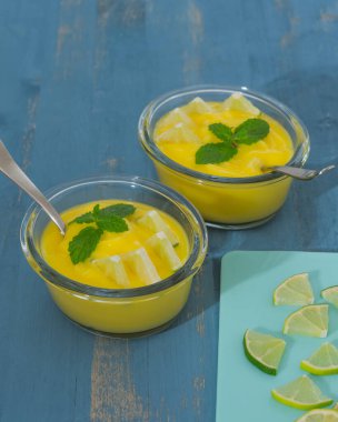 Naneli şeker ve dilimlenmiş limonla servis edilen cam kaselerde limonlu puding mavi ahşap arka planda dikey afiş