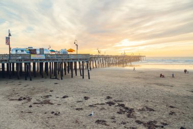 Pismo Plajı, Kaliforniya / ABD - 1 Ocak 2021 Pismo Sahili gün batımı. Geniş kumlu sahil, batan güneşe doğru uzanan ahşap iskele, yürüyen insanlar, ve güzel bulutlu arka plan
