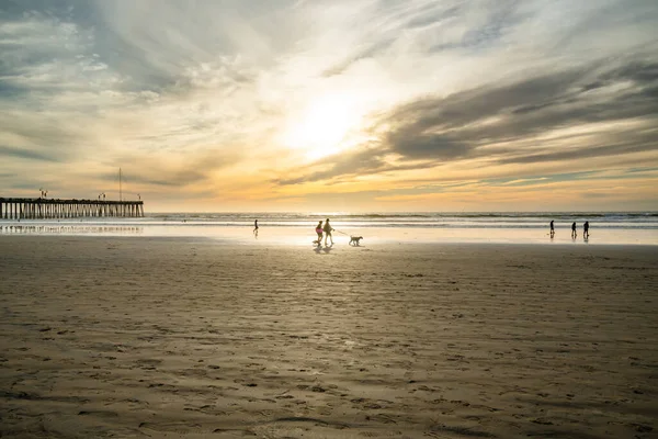 stock image Pismo Beach, California/USA - January 1, 2021 Wide sandy beach at sunset. Walking people, wooden pier, seashore, and sun setting down the horizon, Pismo Beach, California