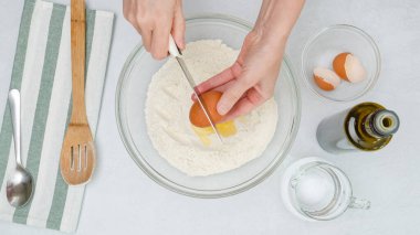 Pişirme malzemelerinin açık gri arkaplanı kapatması gerekiyor. Kadının elleri un dolu bir kaseye yumurta ekler.