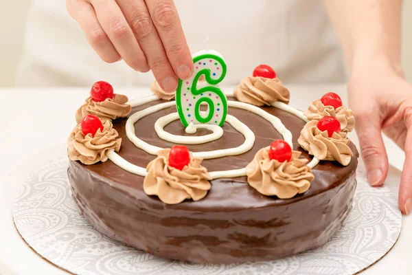 Birthday chocolate cake. Woman hands decorating cake with candle in the form of number Six. Homemade cake decorated with chocolate cream and cherries close up on white background