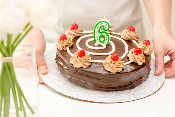 Birthday chocolate cake with candle in the form of number Six in woman hands. Homemade cake decorated with chocolate cream and cherries close up on white background