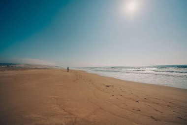 Kumsalda güneşli bir gün, deniz manzarası çok güzel. Turkuaz renkli deniz, geniş kumlu sahil ufukta yürüyen insan silueti ve açık mavi gökyüzü.