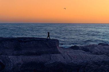 Gün batımı deniz manzarası. Kayalık uçurumlar ve yürüyen kadın silueti, mavi okyanus ve arka planda parlak sarı gökyüzü, kopyalama alanı