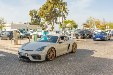 Olhao, Portekiz, 12 Ekim 2025. Gümüş renkli bir Porsche Cayman, marina yakınlarında parlak bir günde renkli spor araba konvoyuna önderlik ediyor..