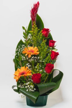Kırmızı güller, turuncu gerberalar ve beyaz arka planda yeşillik dikey çiçek aranjmanı.