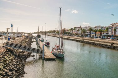 Lagos, Portekiz, 8 Kasım 2025. Sakin bir nehir manzarası. Palmiye ağaçları ve binalarla dolu sessiz bir gezinti güvertesine demirlemiş yelkenliler..