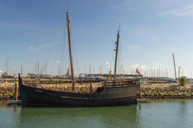 Lagos, Portekiz, 8 Kasım 2025. Geleneksel ahşap bir yelkenli gemi, sakin marina sularında modern yatların ve berrak gökyüzünün arka planında dinleniyor..