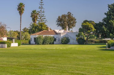 İyi bakımlı yeşil çimenler, yemyeşil ağaçlar ve açık gökyüzü tarafından çerçevelenmiş küçük beyaz bir villaya kadar uzanıyor..