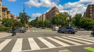 Washington D.C., 24 Mayıs 2025. Meşgul bir şehir yaya geçidi, tarihi mimari ve açık gökyüzü tarafından çerçevelenmiş arabalar, bisikletçiler ve yayaları yakalıyor..
