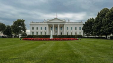 Washington D.C., ABD, 26 Mayıs 2025. Beyaz Saray belirgin bir şekilde çiçek açan bir yatağın ve çeşmenin arkasında duruyor. Amerikan bayrağı, kasvetli bir gökyüzünün altında yarıya indirilmiş..