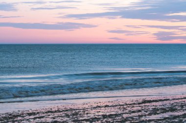 Küçük dalgalar parıldayan pembe ve mor bir sabah gökyüzünün altında kıyıya doğru yuvarlanıyor..