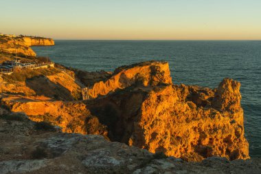 Portekiz, Carvoeiro 'daki kayalıkların altında dalgalar yuvarlanırken engebeli kıyı kayaları altın saat ışığında parlıyor..