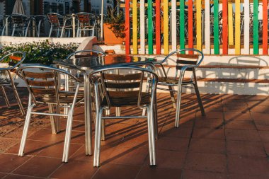 Metal kafe masası ve güneşli terasta parlak boyalı çitlerin yanına yerleştirilmiş sandalyeler..