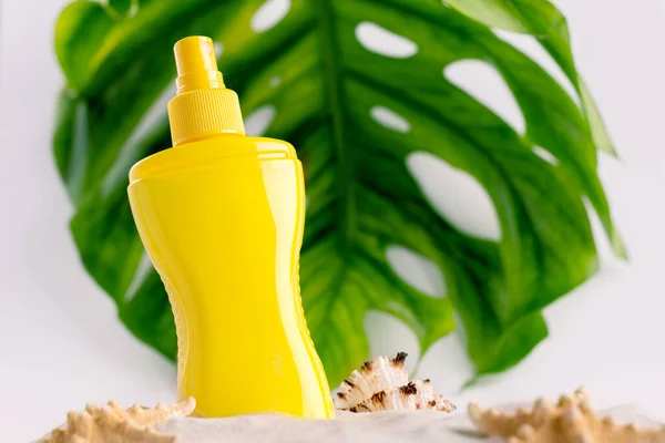 Botella de spray protector solar en una playa de arena con diferentes conchas marinas contra el fondo de la hoja de monstera grande. — Foto de Stock