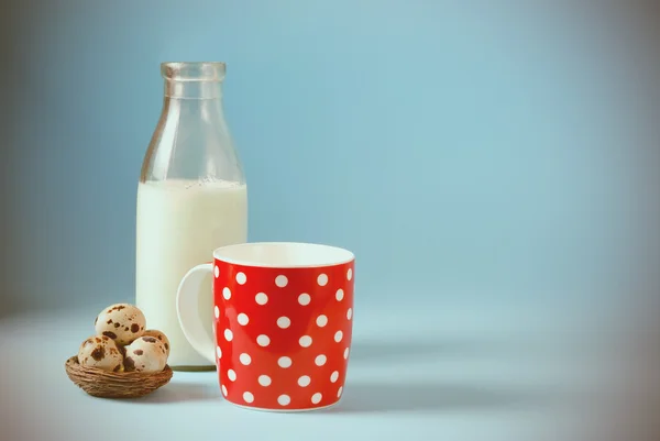 Vintage still life with red, in polka dot, cup of milk, quail eggs, and antique bottle of milk on a blue background. — Stock Photo, Image