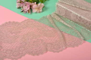 Zarif bej renkli kurdele, mobilya ve çiçekli dantel kumaş, aksesuarlar, bantlar, pembe ve yeşil arka planda yüzükler. Elastik malzeme. Atıcı ve nakış dükkanı için kullanılıyor.