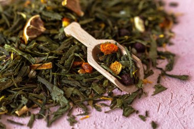 Seçkin yeşil yapraklar Sencha çayı ve şekerli meyveler, şahin üzümü, portakal kabuğu, kuru takviye yaprakları, pembe beton arka planda dondurulmuş deniz dikenleri..