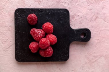 Donmuş taze taze donmuş ahududu yığını. Pembe beton üzerine siyah kesme tahtası. Yaz meyveli vejetaryen konsepti. Seçici odak kapat.