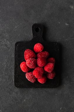Donmuş taze taze donmuş ahududu yığını gri betonun üzerinde siyah kesme tahtasının üzerinde parlak kırmızı ahududu. Yaz meyveli vejetaryen konsepti. Seçici odak kapat.