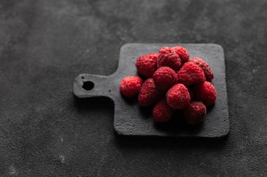 Donmuş taze taze donmuş ahududu yığını gri betonun üzerinde siyah kesme tahtasının üzerinde parlak kırmızı ahududu. Yaz meyveli vejetaryen konsepti. Seçici odak kapat.