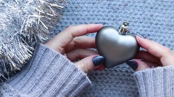 Mani femminili con unghie scintillanti in maglione grigio rotante albero di Natale cuore giocattolo su sfondo lavorato a maglia con ghirlanda di fili di lame d'argento — Video Stock