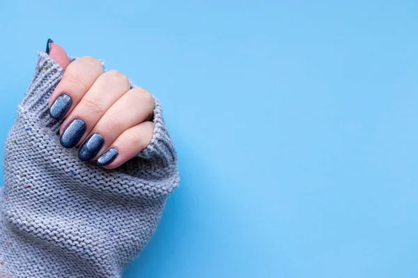 Γυναικείο χέρι σε γκρι πλεκτό ύφασμα πουλόβερ με όμορφο μανικιούρ - σκούρο μπλε γυαλιστερά νύχια σε μπλε φόντο. Επιλεκτική εστίαση. Κλείσιμο προβολής με αντιγραφή χώρου — Φωτογραφία Αρχείου
