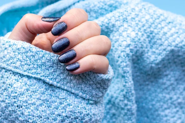 Kvinnlig hand i stickat tröjtyg med vacker manikyr - mörkblå glittrande naglar på blå bakgrund. Selektivt fokus. Närbild — Stockfoto