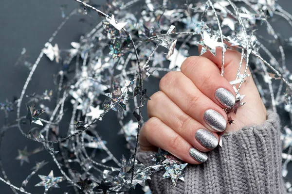 Weibliche Hand in grauem Strickpullover mit schöner Feiertagsmaniküre - silber glitzernde Nägel mit weihnachtlichem Draht und Sternen. Selektiver Fokus. Nahaufnahme. Unscharfer Hintergrund mit Kopierraum — Stockfoto