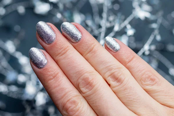 La mano femenina con la manicura hermosa de fiesta - los clavos plateados lustrosos con el alambre retorcido navideño con las estrellas. Enfoque selectivo. Vista de cerca. Fondo gris borroso —  Fotos de Stock