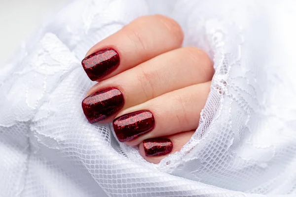 La mano femenina con la manicura hermosa - las uñas oscuras rojas brillantes en la tela blanca de seda con el encaje. Enfoque selectivo —  Fotos de Stock