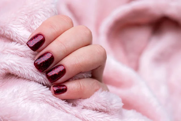 Mano femenina con hermosa manicura - uñas brillantes de color rojo oscuro sobre tela esponjosa rosa, fondo textil con espacio para copiar. Enfoque selectivo —  Fotos de Stock