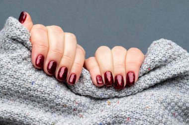 Gri örgü süveter kumaşlı, güzel manikürlü kadın elleri. Gri arka planda koyu kırmızı renkli tırnaklar.