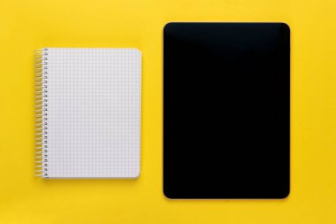Açık spiral kare defter veya boş beyaz kağıtlar ve parlak sarı zemin üzerinde siyah dijital tablet, üst görünüm, düz yatıyordu