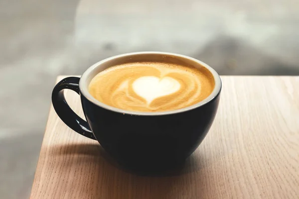 Black cup of coffee with heart shape latte art foam on wooden table in cafe closeup