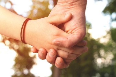 Baba, bulanık doğa arka planına karşı güneş ışığıyla bir kız çocuğunun elini tutuyor. Güven, bakım ve ebeveynlik ailesi, Babalar Günü konsepti