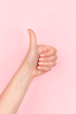 Güzel manikürlü bir kadın eli. Pembe çıplak tırnaklar. Pembe arka planda baş parmak işareti yapıyor.